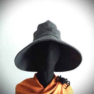 May include: A gray witch hat with a wide brim and a tiered crown. The hat is displayed on a black mannequin head, with an orange scarf and a black spider brooch. The hat is a Halloween costume accessory.