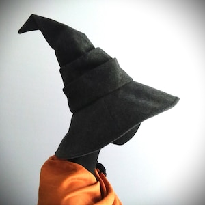 May include: A dark gray witch hat with a wide brim and a pointed top. The hat is made of a soft material and is displayed on a black mannequin head. An orange shawl is draped around the mannequin's neck.