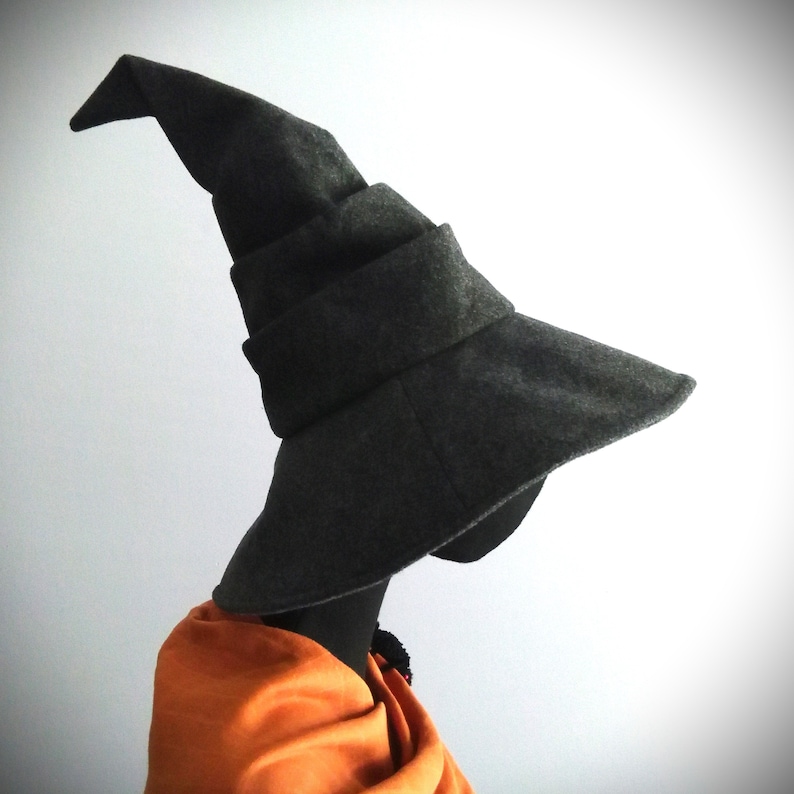 May include: A dark gray witch hat with a wide brim and a pointed top. The hat is made of a soft material and is displayed on a black mannequin head. An orange shawl is draped around the mannequin's neck.