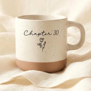 May include: A speckled white ceramic mug with a brown base and handle. The mug features the text "Chapter 30" in script, along with a small floral illustration. The mug is set against a neutral, textured background.