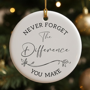 Könnte beinhalten: Ein weißer, runder Weihnachtsschmuck mit dem schwarzen Text "NEVER FORGET The Difference YOU MAKE". Er hängt an einem goldenen Faden, mit einem verschwommenen Weihnachtsbaum und goldenen Kugeln im Hintergrund.