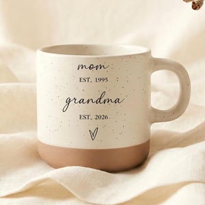 以下が含まれることがあります： ベージュのベースとハンドルが付いた、斑点のある白いセラミックマグ。マグには、筆記体で「mom」と「grandma」の文字と、「EST. 1995」と「EST. 2026」の年号が印刷されています。「grandma」の下には小さなハートが印刷されています。