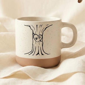 Op de afbeelding: Een witte keramische mok met een bruine basis en een zwart boomontwerp. De boom heeft een hart met de letters "B+D". De mok heeft een oor en is gespikkeld.