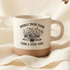 Puede incluir: Una taza de cerámica moteada con base marrón y sección superior blanca. La taza presenta una ilustración negra de un libro abierto con flores y mariposas, y el texto "SPREAD THOSE PAGES LIKE A GOOD GIRL."