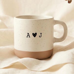 May include: A speckled white ceramic mug with a brown base, featuring the letters "A" and "J" with a heart symbol between them. The mug is set on a cream-colored fabric, with a sprig of dried foliage in the background.