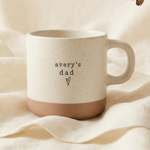 Op de afbeelding: Crèmekleurige keramische mok met een gespikkelde afwerking en een bruine basis. De mok heeft de tekst "avery's dad" met een hartsymbool. De mok staat op crèmekleurige stof.