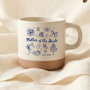 Może przedstawiać: Kremowy ceramiczny kubek z brązową podstawą i uchwytem. Kubek posiada niebieskie ilustracje i napis "Mother of the Bride" z datą. Kubek stoi na kremowej tkaninie.
