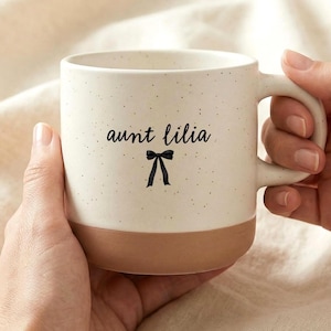 May include: Cream-colored ceramic mug with a speckled finish and a brown base. The mug features the text "aunt lilia" in a script font, with a black bow graphic underneath. The mug is held by two hands.