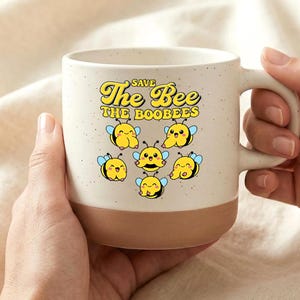 Puede incluir: Taza de cerámica crema y marrón con el texto "Save The Bee The Boobees" y abejas de dibujos animados. La taza está sujeta por dos manos. La taza está diseñada con una parte superior crema y una base marrón.