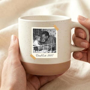 以下が含まれることがあります： 茶色のベースと、父と子の白黒写真が特徴のクリーム色のセラミックマグカップ。写真はテープでマグカップに貼り付けられており、「Dad Est. 1997」の文字が入っています。