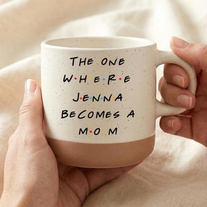 May include: Cream-colored ceramic mug with a brown base, featuring the text "THE ONE WHERE JENNA BECOMES A M.O.M" in black, with the letters "W.H.E.R.E" and "M.O.M" in colorful letters. The mug is held by two hands.