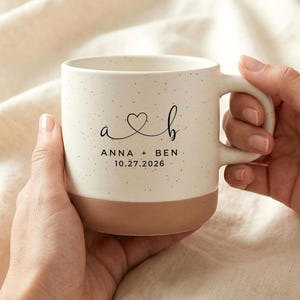 Puede incluir: Taza de cerámica color crema con acabado moteado y base marrón. La taza presenta las iniciales "a" y "b" conectadas por un corazón, junto con los nombres "ANNA + BEN" y la fecha "10.27.2026".