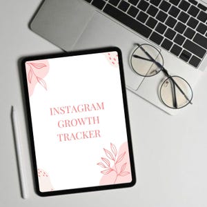 Könnte beinhalten: Ein schwarzes Tablet mit dem Text "INSTAGRAM GROWTH TRACKER" in Rosa, mit rosa floralen Akzenten. Ein weißer Stift, ein Laptop und eine Brille mit runden Gläsern sind ebenfalls auf einer weißen Oberfläche zu sehen.