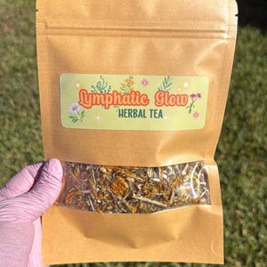 May include: A brown kraft paper pouch containing loose leaf herbal tea. The label features the text "Lymphatic Glow HERBAL TEA" in orange and green, decorated with small floral illustrations and star accents. The bottom section is clear.