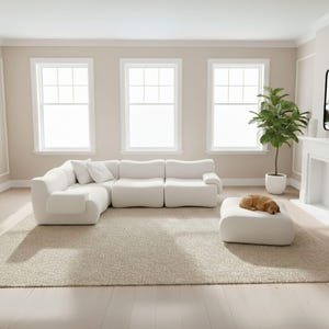 Op de afbeelding: Een moderne, witte hoekbank met bijpassende poef in een neutraal getinte woonkamer. De bank heeft een gebogen ontwerp en wordt gecombineerd met een beige tapijt. Een kleine hond rust op de poef. De kamer heeft drie grote ramen en een plant in een pot.