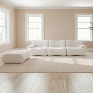 May include: A modern living room featuring a white modular sofa and matching ottoman. The room has beige walls, hardwood floors, and a patterned rug. Large windows provide natural light, creating a bright and airy atmosphere.