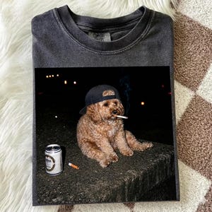 May include: A charcoal gray crewneck t-shirt featuring a graphic print of a light brown curly-haired dog wearing a black baseball cap, holding a cigarette in its mouth, sitting next to a silver beverage can and a discarded cigarette on the ground.