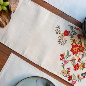 May include: Cream-colored table runner with embroidered floral design in red, yellow, and green. The runner is rectangular and placed on a wooden table. A matching placemat and blue plates are also visible.