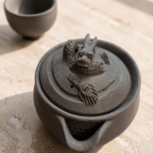 Op de afbeelding: Een donkergrijze, matte keramische theepot met een deksel voorzien van een gebeeldhouwde drakenfiguur en een ingebouwde zeef bij de tuit, vergezeld door een bijpassend klein keramisch kopje. De set heeft een textuurrijke, aardse afwerking.