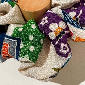 Op de afbeelding: Een houten flesstopper omringd door kleurrijke stukken stof. De stof heeft patronen in groen, paars, rood en blauw. De stopper is cilindrisch en gemaakt van lichtgekleurd hout. De stukken stof zijn gevouwen en rond de stopper gerangschikt.