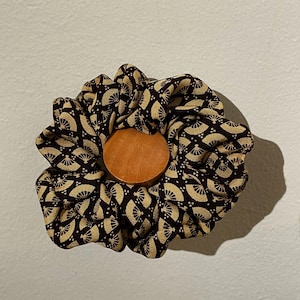 Op de afbeelding: Een scrunchie met een bruine en gouden waaierpatroonstof, gecentreerd met een ronde houten knoop. De stof is donkerbruin met een herhalend gouden waaierontwerp. Een haaraccessoire.