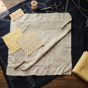 Può includere: Una collezione di campioni di tessuto e accessori per cucire su una superficie di legno. I campioni includono tessuti fantasia e strutturati in beige e giallo. Sono presenti anche un tessuto blu navy con cuciture bianche, forbici e una bobina di filo. Una piccola etichetta recita "Renaras".