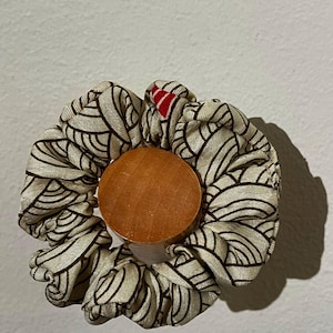 Op de afbeelding: Een crèmekleurige scrunchie met een zwart, golfachtig patroon en een klein rood accent. Het is gecentreerd rond een rond houten object. Dit accessoire wordt gebruikt om haar vast te binden.