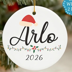 Könnte beinhalten: Ein weißer Keramik-Weihnachtsschmuck mit dem Namen "Arlo" in schwarzer Schrift, bedeckt mit einem Weihnachtsmannhut. Das Jahr "2026" steht unter dem Namen, mit einer festlichen Girlande. Der Schmuck hängt an einer goldenen Schnur.