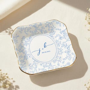 Op de afbeelding: Een vierkant, wit keramisch sieradenschaaltje met een blauw bloemmotief en gouden rand. Het midden heeft een witte cirkel met de initialen "j + k" en de datum "09.20.2025" in blauw schrift. Ideaal voor bruilofts- of jubileumcadeaus.