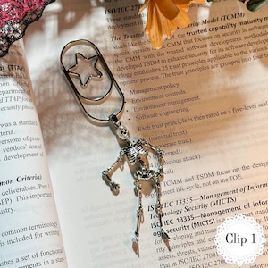 May include: Silver-toned metal bookmark featuring a star-shaped cutout and a dangling skeleton charm. The bookmark is attached to a paper with text. The text "Clip 1" is in the bottom right corner.