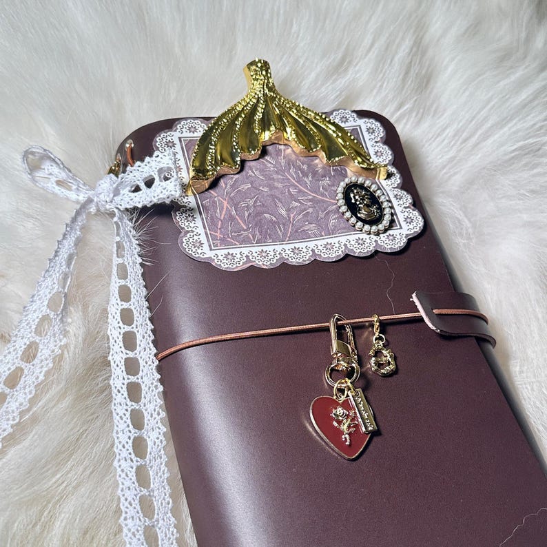 May include: A dark brown leather journal with a gold-colored decorative accent and a white lace ribbon. The journal features a heart-shaped charm and a small gold-colored charm. The journal is closed with a brown leather strap.