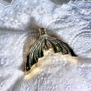 May include: A gold-coloured mermaid tail-shaped hair clip. The clip has a detailed, textured design with a fan-like shape. The clip is lying on a white, embroidered fabric.