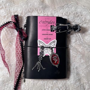 May include: Black leather journal with a pink label that reads "everyday." The journal is adorned with a silver hand-shaped clip, a butterfly sticker, and a red rose pendant. A black string wraps around the journal. A ribbon with lace and polka dots is on the left.