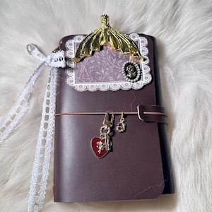 May include: A dark brown leather journal with a gold-colored decorative accent and a red heart charm. The journal has a lace ribbon and a decorative frame with a cameo. The journal is closed with a brown elastic band.