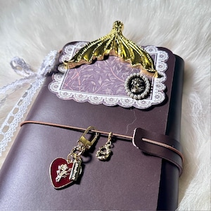 May include: A dark purple journal with a gold-colored decorative clip and a heart-shaped charm. The journal has a decorative lace trim and a brown leather strap. The heart charm has a rose design.