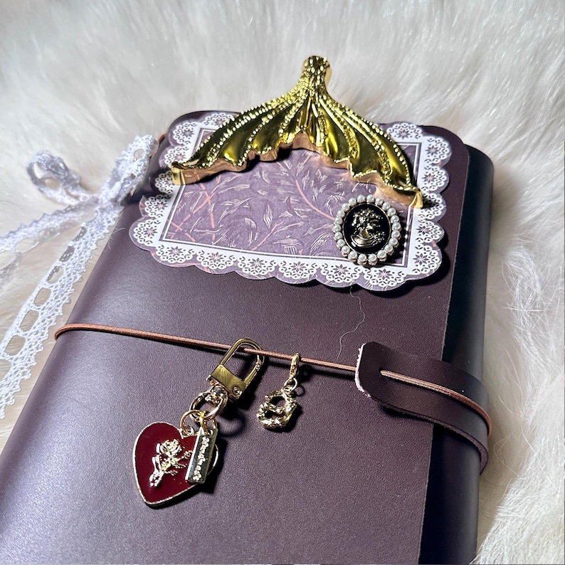 May include: A dark purple journal with a gold-colored decorative clip and a heart-shaped charm. The journal has a decorative lace trim and a brown leather strap. The heart charm has a rose design.