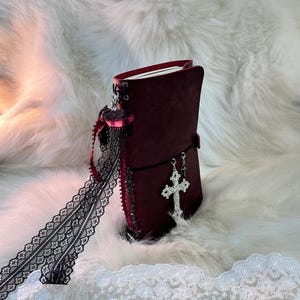 May include: A burgundy leather journal with a black elastic closure and a silver cross charm. The journal is decorated with black lace and a burgundy ribbon. The pages are visible inside the journal.