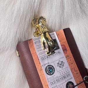 May include: A gold-coloured hand-shaped paper clip securing a vintage ticket to a brown leather notebook. The ticket features printed text and numbers, including "OLD 09.48.1946" and "OLD 98.4196.64". The notebook rests on a white, fluffy surface.
