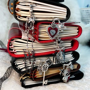 Puede incluir: Una pila de diarios encuadernados en cuero negro y rojo, adornados con colgantes decorativos plateados. Los colgantes incluyen un esqueleto, un corazón con un ojo, llaves y otros diseños de inspiración gótica. Los diarios tienen páginas color crema.