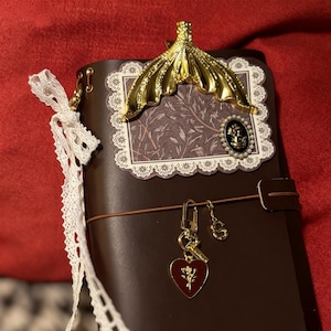 May include: A brown leather journal with a gold-colored decorative accent and a decorative paper insert. The journal features a gold charm with a heart and key, and a white lace ribbon.