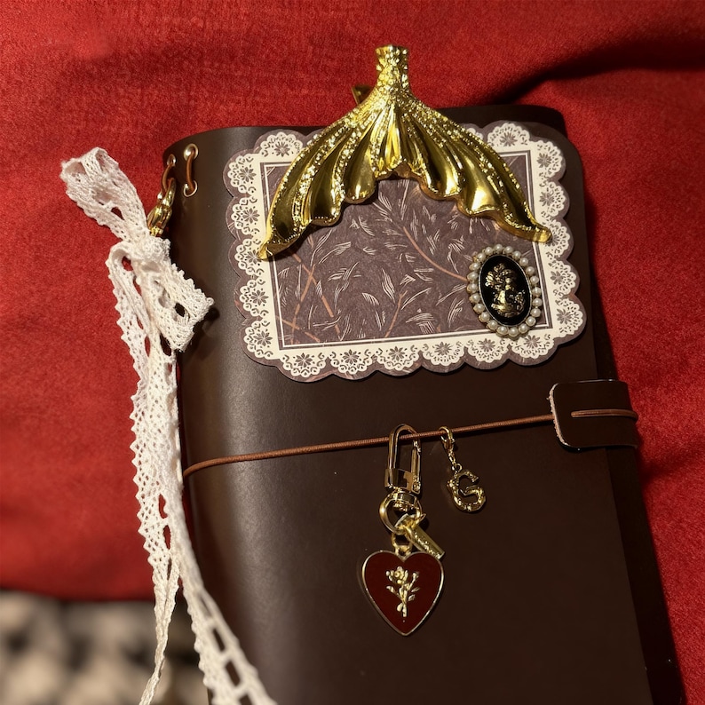 May include: A brown leather journal with a gold-colored decorative accent and a decorative paper insert. The journal features a gold charm with a heart and key, and a white lace ribbon.