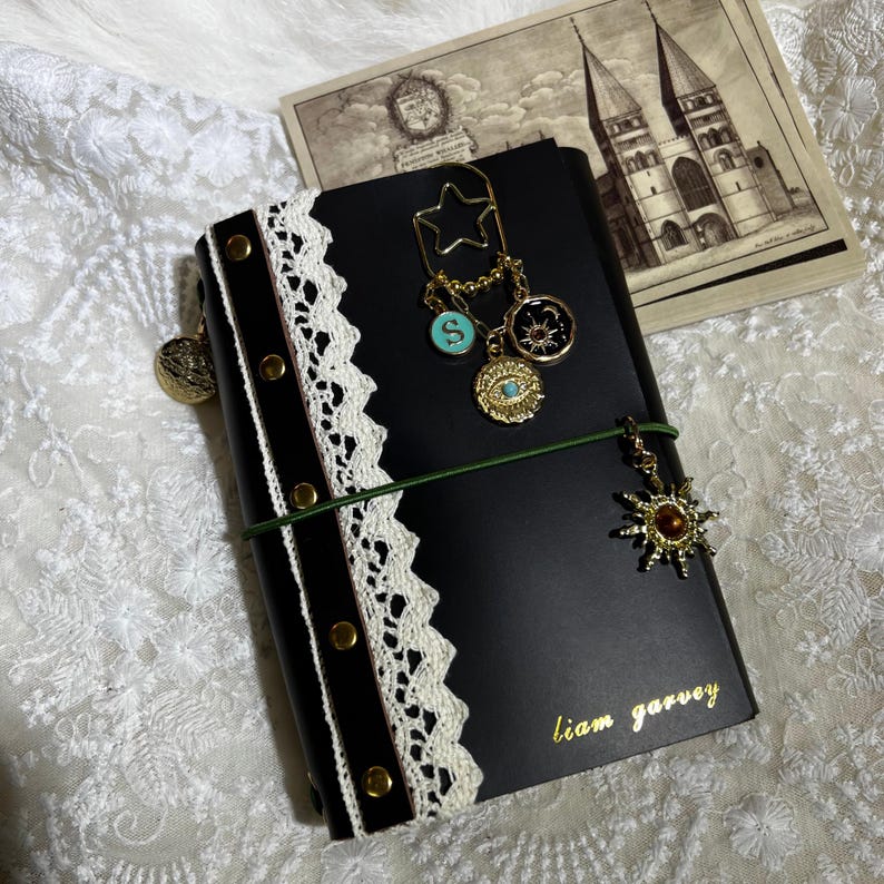 Puede incluir: Un cuaderno negro con borde de encaje blanco, tachuelas doradas y el nombre liam garvey impreso en oro. Est&aacute; adornado con un clip en forma de estrella, un dije turquesa con la letra S, un dije de sol y luna, y un dije de sol dorado en una banda verde.