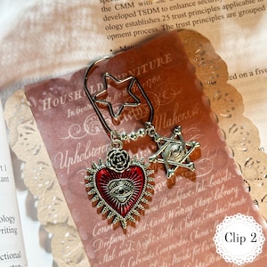 May include: A decorative metal bookmark with a star-shaped clip. It features a red heart pendant with an eye design and a six-pointed star pendant. The bookmark is on a vintage paper background.