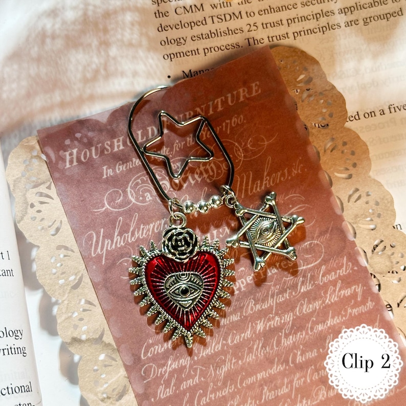 May include: A decorative metal bookmark with a star-shaped clip. It features a red heart pendant with an eye design and a six-pointed star pendant. The bookmark is on a vintage paper background.