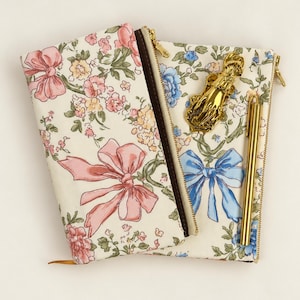 May include: Two zippered pouches made of cream-colored fabric with a floral pattern featuring pink and blue bows. A gold-toned metal hand-shaped clip and a slim gold-toned metal pen are placed on top of the right pouch.