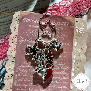 May include: A silver bookmark with a star-shaped top and dangling charms. Charms include a black star, a red and silver heart with wings, and a silver heart with wings. The bookmark is displayed on a vintage paper background.