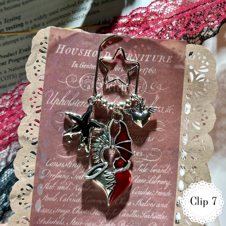 May include: A silver bookmark with a star-shaped top and dangling charms. Charms include a black star, a red and silver heart with wings, and a silver heart with wings. The bookmark is displayed on a vintage paper background.