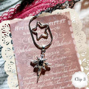 May include: A silver-colored metal bookmark with a star-shaped top and a starburst pendant with a black center stone. The bookmark is clipped to a pink paper with text.