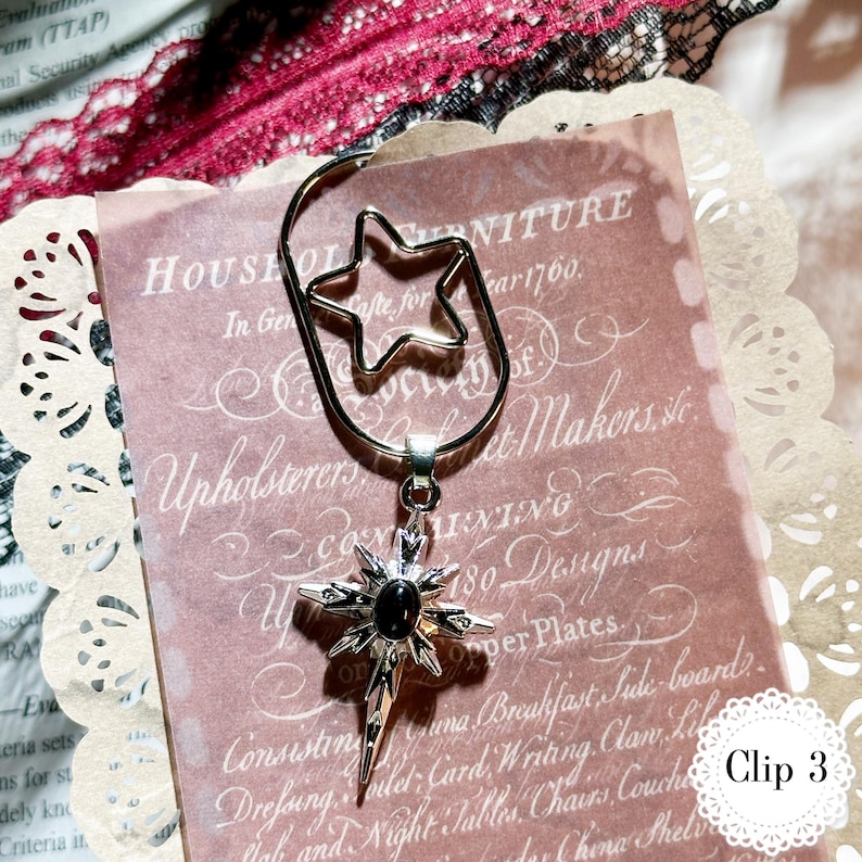 May include: A silver-colored metal bookmark with a star-shaped top and a starburst pendant with a black center stone. The bookmark is clipped to a pink paper with text.