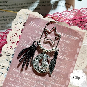 May include: A metal bookmark with a star-shaped top and decorative charms. Charms include a black skeleton hand, a crescent moon with a wolf, and a knife. The bookmark is on a pink paper with text.